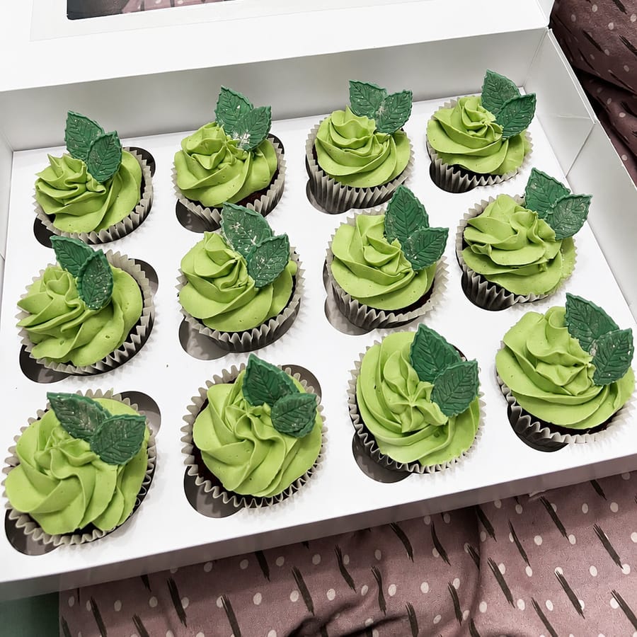 A boxed dozen of chocolate cupcakes with green buttercream swirls and delicate sugar leaves