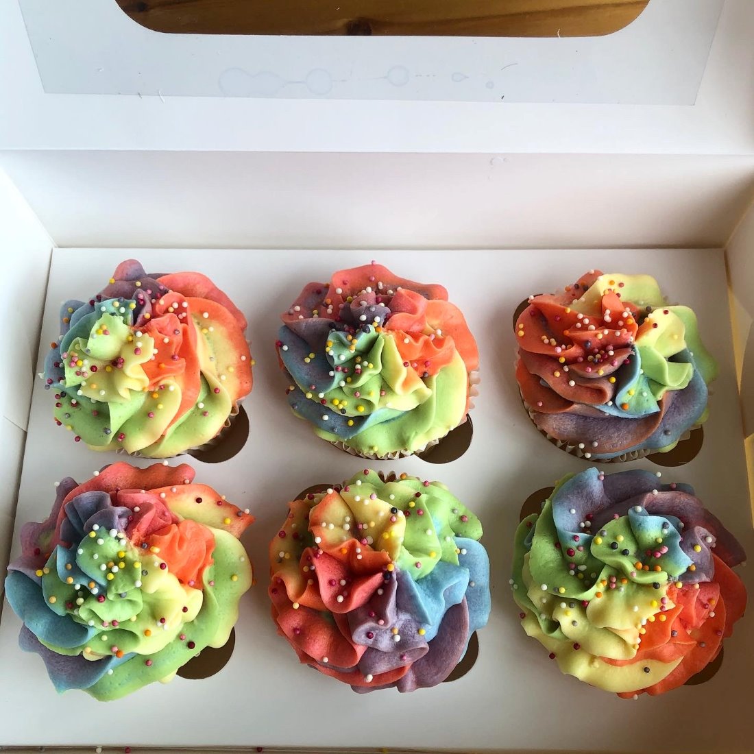 A box of six vanilla cupcakes piped with rainbow swirl buttercream and sprinkles